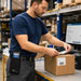 Warehouse worker wearing an AgozTech mobile computer case while packing a labeled box at a logistics workstation.