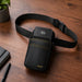 A black AGOZ POS waist bag with a built-in black rectangular device, shown on a wooden tabletop with a plant and notebook in the background.