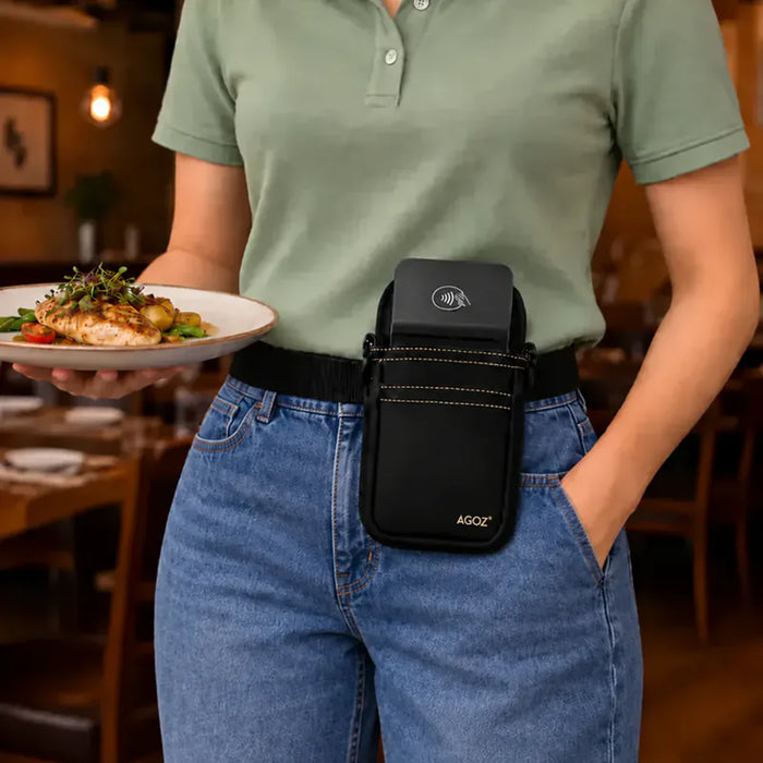 A person in a restaurant setting wearing a black waist bag with a Adyen POS holder, holding a plate of food in one hand and wearing blue jeans in the other. The bag has a black neoprene exterior, a black belt, and a black phone case insert.