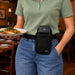 A person in a restaurant setting wearing a black waist bag with a Adyen POS holder, holding a plate of food in one hand and wearing blue jeans in the other. The bag has a black neoprene exterior, a black belt, and a black phone case insert.