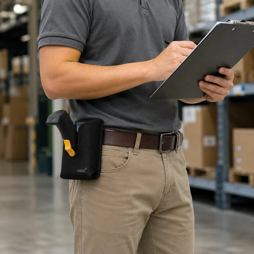 A person in a gray polo shirt and khaki pants stands in a warehouse holding a clipboard, with a black Bluebird scanner holster containing a grip attached at the waist.